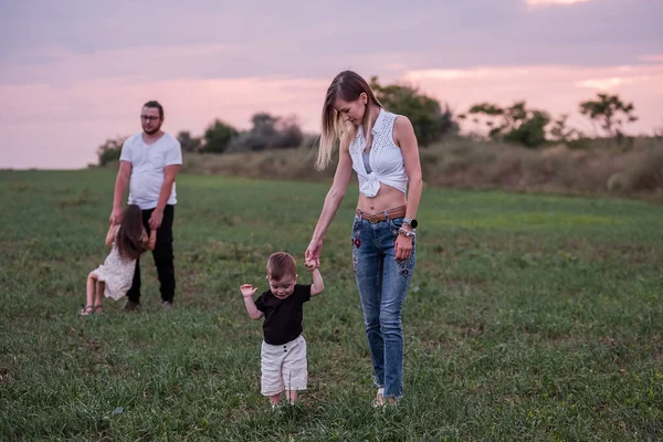 Woman helps toddler walk in a grassy field at sunset, with man and child in the background, capturing a casual, loving diversity family moment in the countryside. First steps