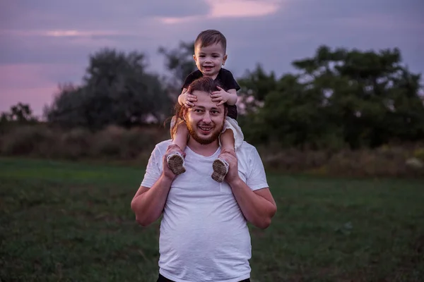 Diversity father with beard carrying son on shoulders shares tender moment in twilight lit field, embodying the essence of paternal love. Man and boy fooling around, playing. Child touches mans nose