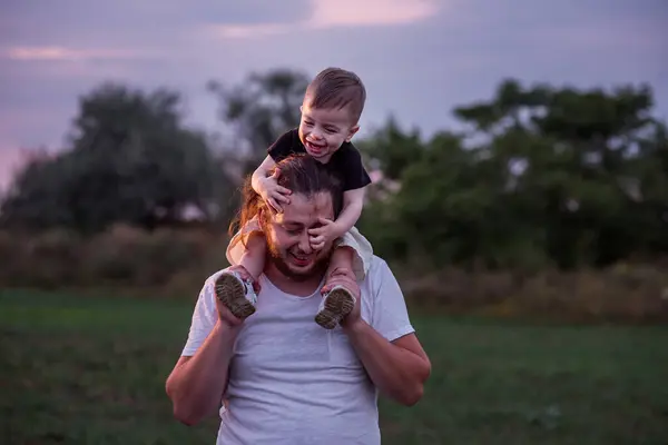 Diversity father with beard carrying son on shoulders shares tender moment in twilight lit field, embodying the essence of paternal love. Man and boy fooling around, playing. Child touches mans nose