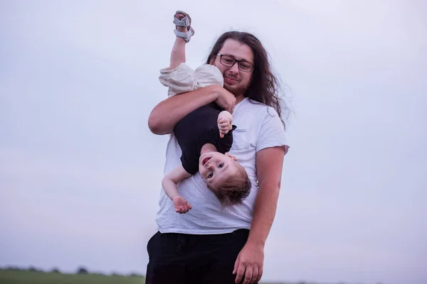 Diversity father holding little son upside down playfully indicates his joyous child. Young man with long curly hair, beard, glasses fooling around with toddler boy on sunset green field