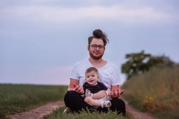 Seated on tranquil path, young, diversity father with happy son on lap, both enjoying peaceful twilight atmosphere in countryside. Man with hair bun, beard playing with funny boy