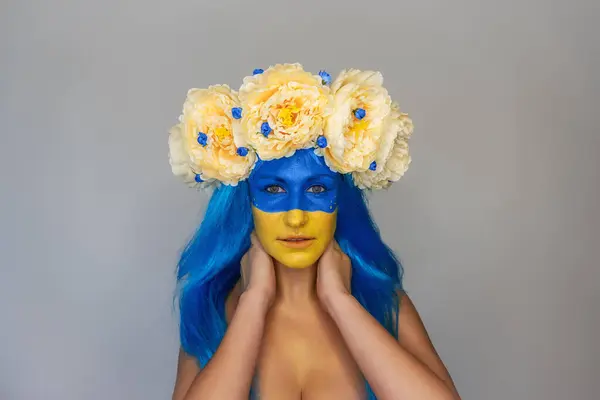 Creative close-up portrait of young Ukrainian patriot woman with yellow blue face art, wreath of flowers on head on isolated gray background. Flag Day, Independence Day in Ukraine. Copy space