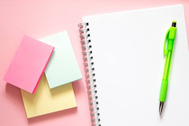 Okula dönüş kavramı. Hassas pembe arka planda üç tane renkli çıkartma. Sirchu 'ya sarmal ve yeşil kalemli beyaz bir defter. Metin için yer alan düzen ve düz uzanma.