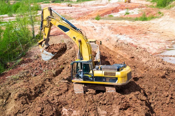 Red clay mining. In a quarry, a yellow excavator with a large bucket loads clay into a truck.