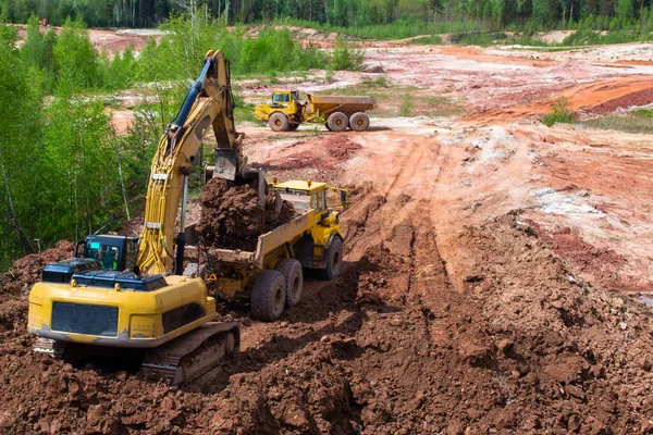 A escavadora amarela cava argila vermelha com um balde grande e coloca-a no caminhão. Mineração de argila . — Fotografia de Stock