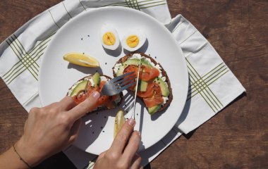 Krem peynirli, avokadolu, balıklı, yumurtalı tahıllı sandviçlerden oluşan sağlıklı bir kahvaltı yapan genç bir kadının elleri. Kahverengi ahşap arka plan.