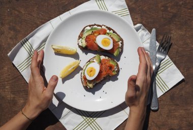 Sağlıklı kahvaltı. Beyaz bir tabak dolusu tahıllı sandviç, krem peynir, avokado, balık ve yumurta tutan genç bir kadının elleri. Kahverengi ahşap arka plan.
