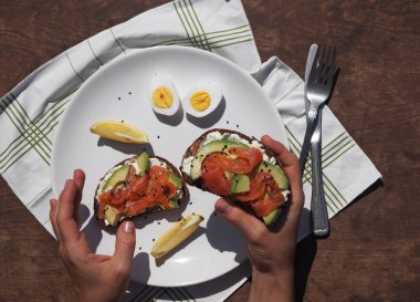 Sağlıklı, doğal yaz yemekleri. Genç bir kadının elleri krem peynirli, avokadolu, balıklı, yumurtalı tahıllı sandviçler hazırlıyor. Kahverengi ahşap arka plan.