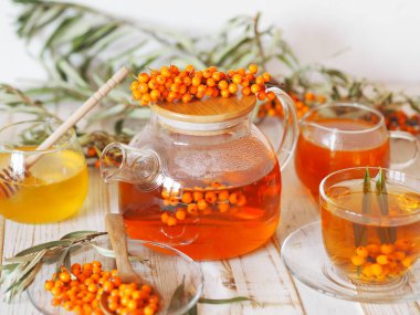 Deniz dikenli çay ile tıbbi arka plan. Çay ile cam çaydanlık, deniz dikenleri, bal ve reçel beyaz ahşap bir masada..