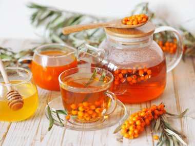 Cam bardakta vitamin dolu deniz dikenli çay çaydanlık, bal, reçel, beyaz bir mutfak masasında taze çiğ deniz dikenli böğürtlen ve filizler. Deniz dikenli üzüm toplama mevsimi..