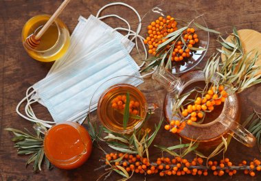 Deniz dikenli dallı tıbbi maskeler, çaydanlık, bal, reçel, ahşap zemin üzerinde taze çiğ deniz dikenli üzümleri ve dalları olan çay. Gribi sağlıklı deniz dikenli böğürtlenlerle koruma ve tedavi etme fikri.