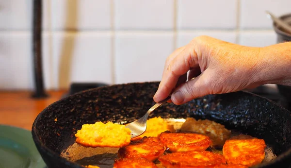 An elderly woman prepares natural diet pancakes from pumpkin and millet in a frying pan on the stove. Home cooking, rustic style. — Stock Image Elderly Woman Prepares Natural Diet Pancakes Pumpkin Millet Frying Pan Stock Picture
