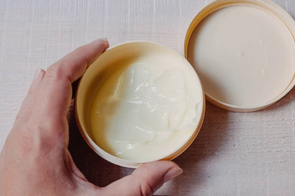 Skin and body care. Open beige jar with cream for the body, hands and feet in a woman\'s hand on a natural white wooden background close-up with soft focus. Photo in the style of fine art.