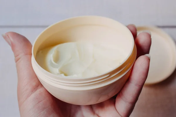 Skin and body care. Open beige jar with cream for the body, hands and feet in a woman's hand on a natural white wooden background close-up with soft focus. Photo in the style of fine art.