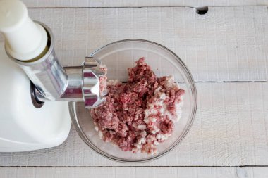 Elektrikli et öğütücüsünde kıyma yapmak. Kıymalı cam kase ve elektrikli et öğütücüsünden elde edilen kıyma beyaz ahşap arka planda yakın plan, üst manzara.