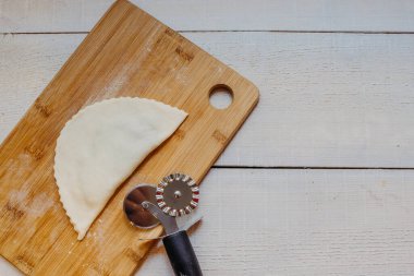 Rus mutfağı. Evde cheburek pişiriyorum. Kızartma çeburek ve patenli bıçak için hazır. Beyaz ahşap arka planda kopi alanı olan tahta bir tahtada hamurun kenarını kesmek için..