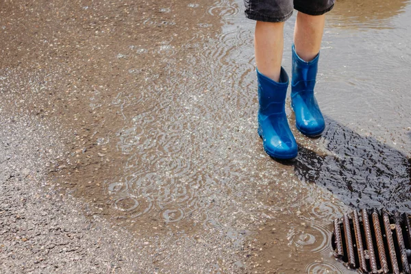Children in the summer after the rain. Close-up of a boy\'s feet in blue rubber boots standing in a puddle on a walk. Children walk in the open air after the rain. Waterproof shoes for boys.