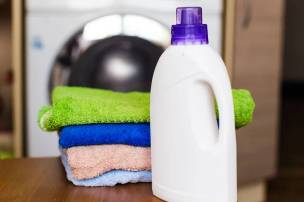 Washing towels in the washing machine. A stack of colorful Terry towels on a wooden table and a white bottle of Laundry detergent in close-up with a washing machine in the blurry background.