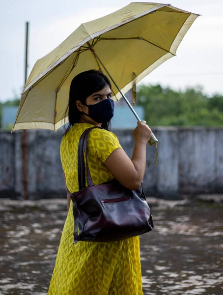 an Indian woman in yellow dress and face mask on is going outside with umbrella and bag after unlock 1.0 of corona virus pandemic