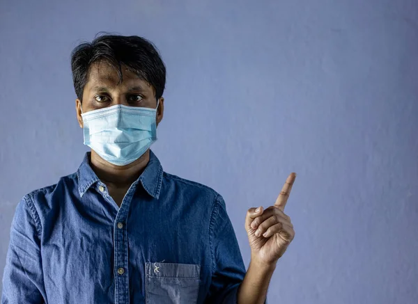 an Indian man wearing surgical nose mask, looking at camera and pointing left side for corona virus or covid-19 protection on white background