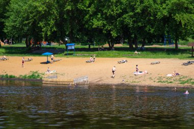 İnsanlar yazın nehir kıyısında dinlenirler. Yeşil ağaçların arka planındaki danslarda güneşlenirler
