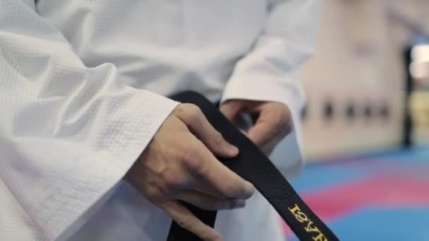 Muž v bílých karate šatech s černým opaskem v pase, zapíná černý opasek — Stock video