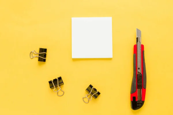 White square notes, red office knife, bulldog paper clip on yellow background with copy space. Mock up. Top view. Concept chancellery, office supplies, accessories