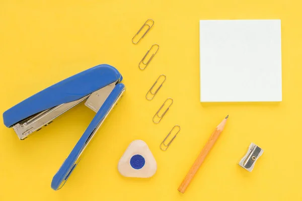 White square notes, pencil, eraser, blue stapler, sharpener, metal paper clip on yellow paper background