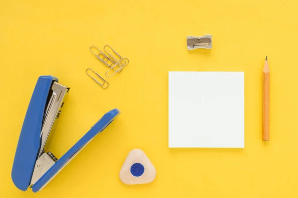 White square notes, pencil, eraser, blue stapler, sharpener, metal paper clip on yellow paper background