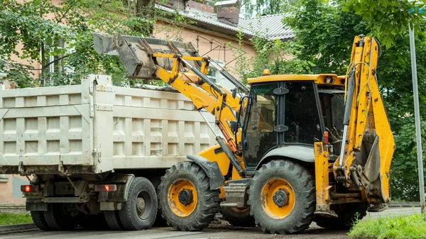 Sarı traktör bir çöp kamyonunun arkasına inşaat enkazı döküyor.