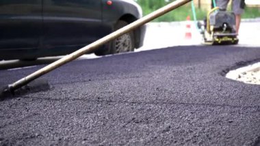 Yakından, yolda ve kaldırımda asfalt döşemek için siyah çakıl küreyen işçiler, dışarıda çalışmak, sıkı çalışmak. Elle titreşen bir silindirle asfalt sermek.