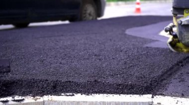 Yakından, yolda ve kaldırımda asfalt döşemek için siyah çakıl küreyen işçiler, dışarıda çalışmak, sıkı çalışmak. Elle titreşen bir silindirle asfalt sermek.