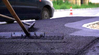 Yakından, yolda ve kaldırımda asfalt döşemek için siyah çakıl küreyen işçiler, dışarıda çalışmak, sıkı çalışmak. Elle titreşen bir silindirle asfalt sermek.
