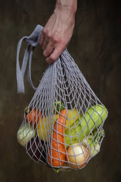 Woman holds her hand gray bag string bag with vegetables,zucchini,bell peppers,carrots,beets,onions brown wooden background.Harvest from garden fresh bright vegetables.Concept zero waste.