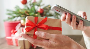 Elinde hediye kutusu ve akıllı telefon olan genç bir kadın Noel dekoru ve hediyelerin arka planında, yakın plan. İnternetten Noel ve Yeni Yıl alışverişi, kredi kartıyla ödeme.