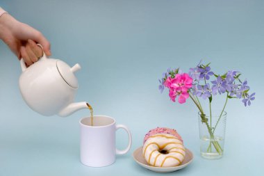 Bir kadının eli, beyaz bir çaydanlıktan beyaz bir fincana, iki çörekli bir tabağa ve bir vazo mavi çiçeğe çay dolduruyor. Fotoğraf mavi bir arkaplanda pastel renklerle yapılır. Kahvaltı konsepti..