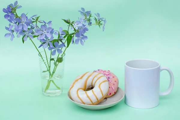 White mug without logos on mint background. There are 2 delicious doughnuts on the plate. There are blue flowers in the vase. Photos in pastel colors. The concept of the morning coffee, Breakfast