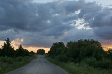 Yol kasvetli, korkunç bulutların altında gidiyor. Uzakta altın bir günbatımı. Yatay fotoğraf