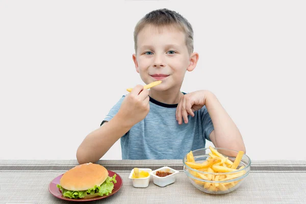 Küçük bir anaokulu öğrencisi masada oturuyor ve ellerinde patates kızartması tutuyor. Fast food, hamburger, patates kızartması ve sos masada. Çocuk büyüyen vücuda zararlı yiyecekler yiyor.