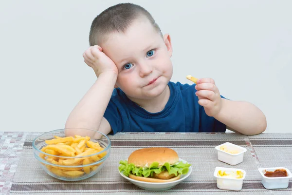 Küçük mavi gözlü çocuk fast food yiyor. Hafif bir arka planda, çocuk üzgün, gülümsemiyor. Kavram - çocuk sağlıksız, monoton yiyeceklerden, lezzetli, sağlıklı ve sağlıklı yiyeceklerin hayallerinden bıktı.