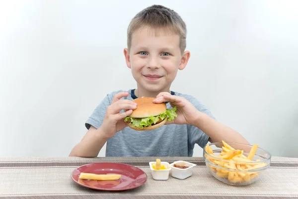 Çocuk masada oturuyor ve elinde taze bir hamburger tutuyor. Gülümsüyor, masada hazır yemek var. Sağlıklı beslenme getirmeyen ama çocuklar arasında çok popüler bir konsept.