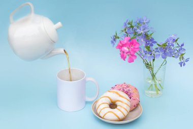 Beyaz çaydanlıktan logoları olmayan bir bardağa sıcak bir içecek dökülüyor. Masanın üzerinde iki güzel kokulu çörek ve bir buket mavi ve parlak pembe çiçekler var. Pastel renklerde fotoğraf, insanlar olmadan.