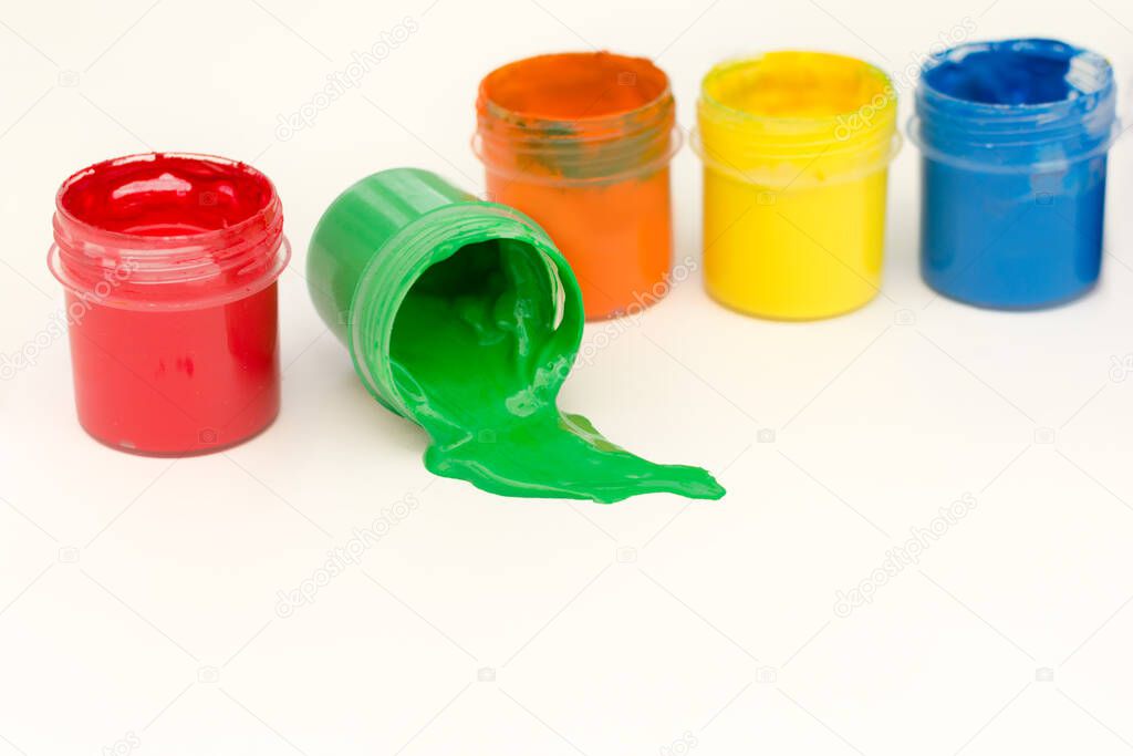 multicolored cans of gouache stand in a row on a white background, one may have fallen and green paint spilled out of it. Horizontal photo, close-up