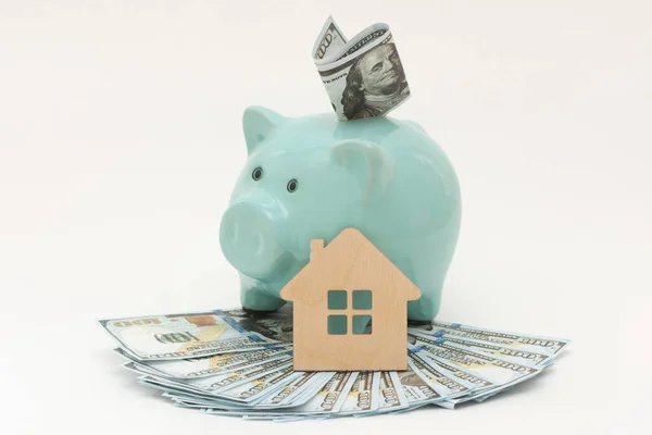 a sparkling new piggy bank sits on a fan of 100 bills,a bill inserted in a piggy bank,next to a wooden house.The idea of buying real estate,affordable housing,loans, mortgages, making money on housing