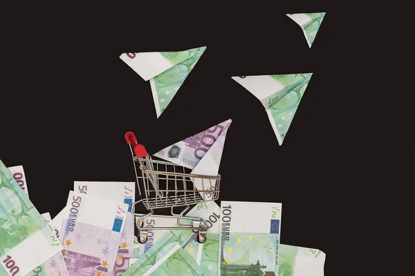 against a black background, a mini-cart from a supermarket stands on money, and planes from euro bills seem to fly out of it. Idea-cost overruns, loss of money, investment,investment of money,profit