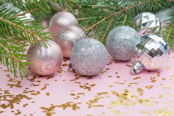 Silver and pink Christmas balls lie on a pastel pink background strewn with golden confetti and fir branches. Photo close-up, horizontal. Concept - layout for a New Year\'s card