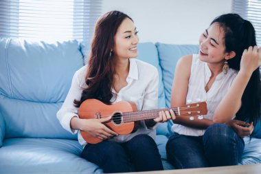 İki Asyalı kadın ukulele çalarak ve evde gülümseyerek eğleniyorlar.