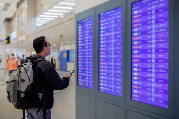 亚洲男子背包旅客使用智能手机在现代机场的航班信息屏幕办理登机手续 — 图库照片