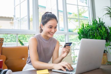 Asyalı bir kadın kredi kartı tutuyor ve evde akıllı telefon alışverişi yapıyor. Çevrimiçi alışveriş, e-ticaret, internet bankacılığı, harcama, ev konseptinden çalışma