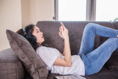 Asyalı, mutlu, neşeli, güzel bir kadın cep telefonundan müzik dinlemek için kulaklık takıyor ve akıllı telefon çalıyor ve evdeki kanepede uzanıyor., 